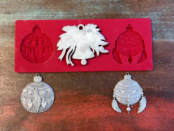 A 14 second video of Zuri Design's Bohemian Christmas Ornaments silicone mold and silver colored castings are against a wood background. A hand is shown holding one of the ornament castings.