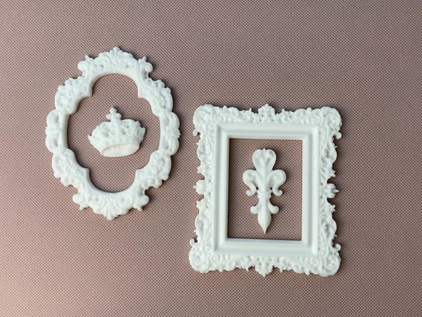 A hand is shown picking up and setting down white resin castings of 2 ornate frames, a small royal crown, and a fleur de lis to show their size against a light beige background.