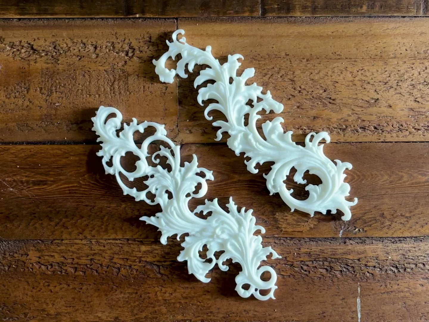 A 12 second video shows a close-up of 2 white resin castings of Baroque Flourishes against a dark wood background. A hand is shown picking up and holding 1 of the castings.