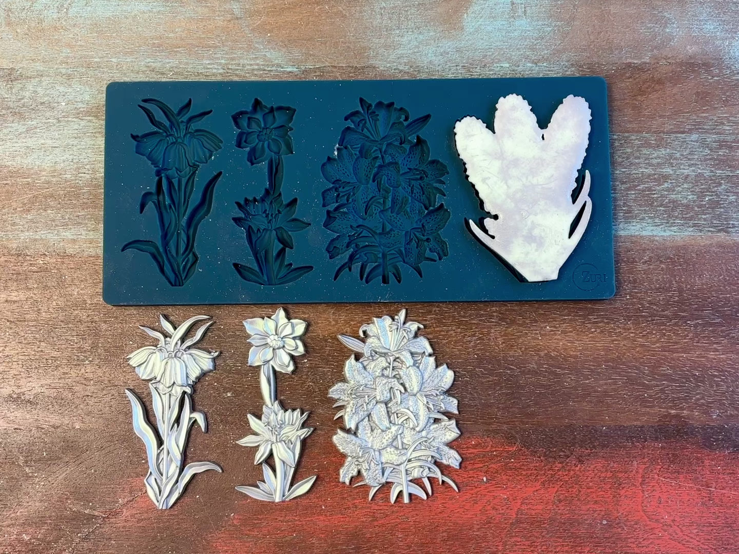 A 13 second video of Zuri Design's Flower Blooms Set 2 are against a wood background. A hand is shown holding one of the castings.