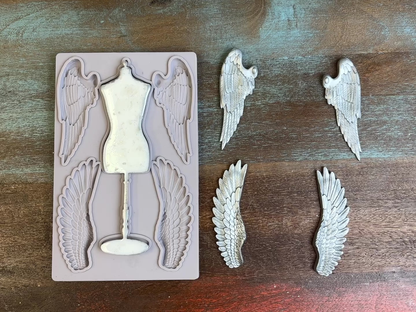 A 14 second video of a light grey silicone mold and silver colored castings of a seamstress dress form and 2 sets of angel wings are against a dark wood background. A hand is shown picking up and flipping over the bust casting to show the details.