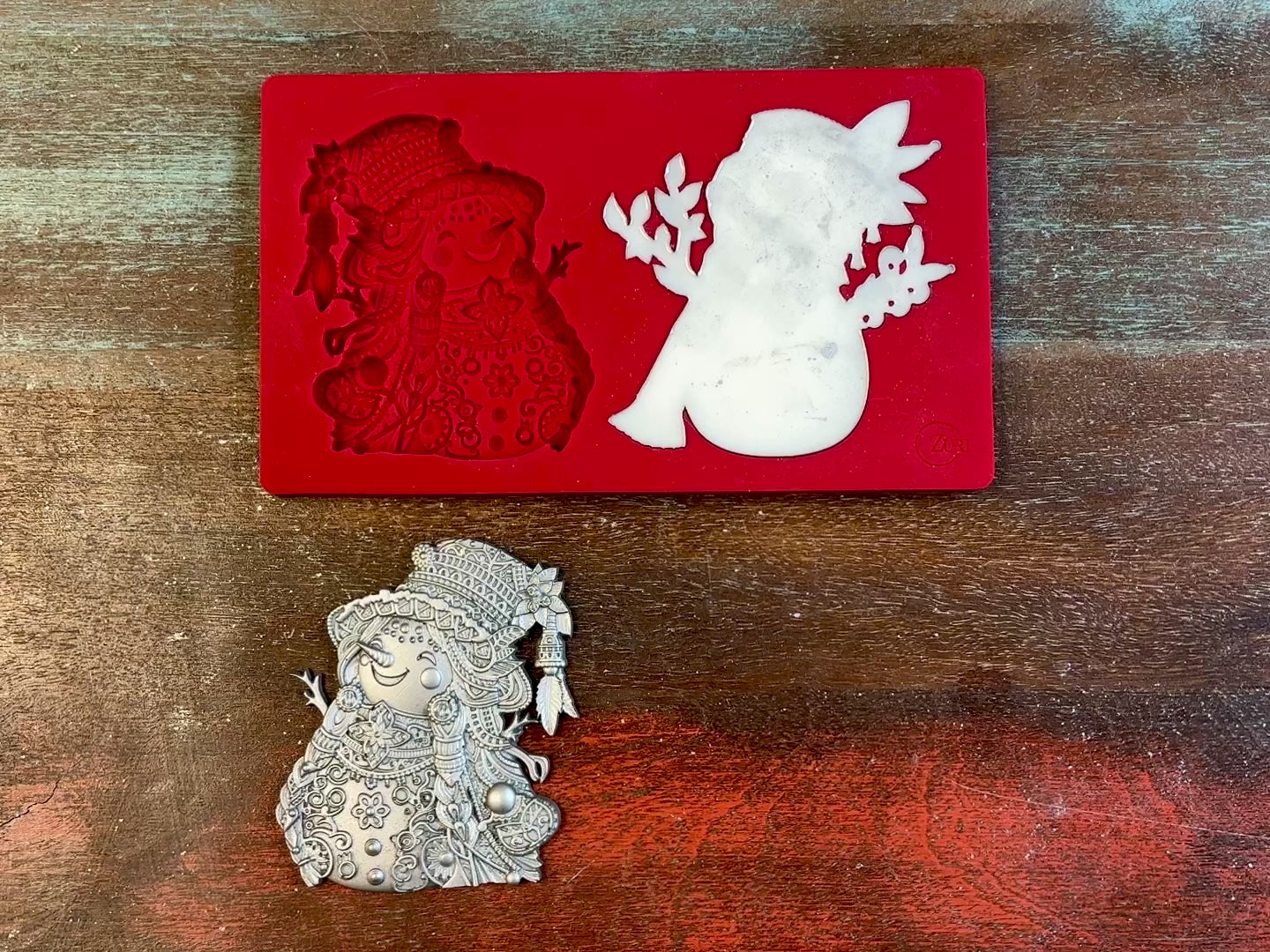 A 14 second video of Zuri Design's Snowman Duo silicone mold and silver colored castings are against a wood background. A hand is shown holding one of two snowman castings.