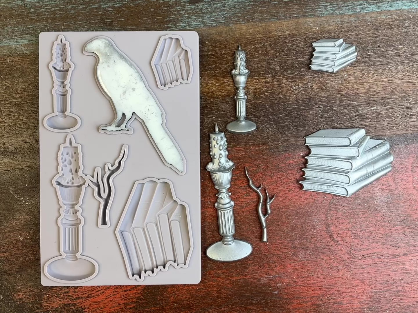 A 15 second video of a light grey silicone mold and silver colored castings of stacks of books, candles, a branch, and a bird are against a dark wood background. A hand is shown picking up and flipping over the bird casting to show the details.