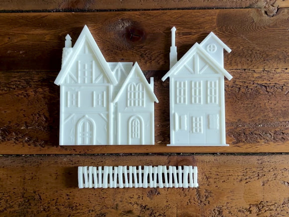 A 14 second video shows a close-up of white resin castings of 2 houses and a picket fence against a dark wood background. A hand is shown picking up one of the houses.