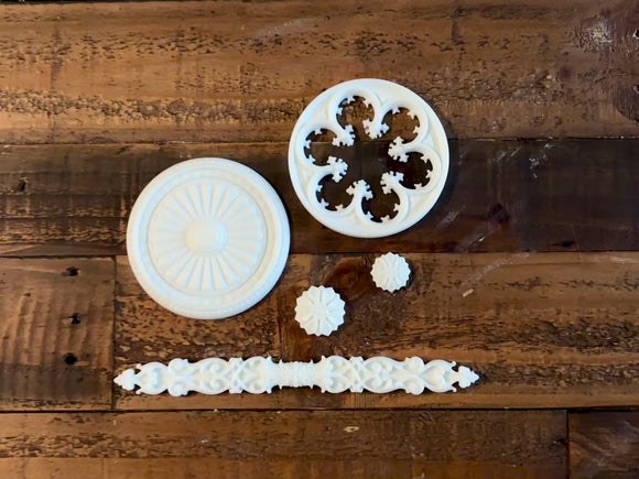 An 11 second video shows a close-up and backside of 5 white resin castings of round ornate windows, accents medallions, and a decorative rod against a wood background. A hand is shown picking up one of the large window castings.