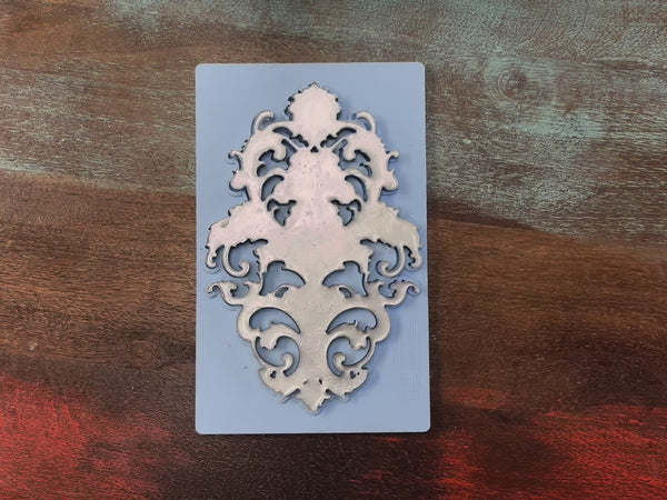 A 13 second video shows a hand lifting and setting down Lablanche's Baroque Emblem silicone mold and a silver colored casting from it against a wood background.