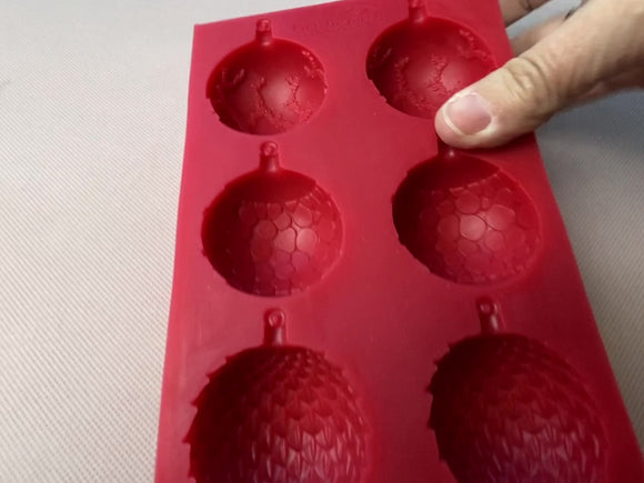 A 15 second video shows a red silicone mold that features 6 half ornate ornament bulbs. A hand places 3 white resin castings of the bulbs on the red mold.