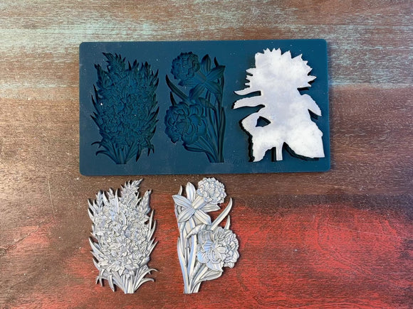 A 13 second video of Zuri Design's Flower Blooms Set 1 silicone mold and silver colored castings are against a wood background. A hand is shown holding one of the castings.