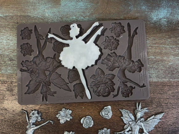 A 15 second video of a brown silicone mold and silver colored castings of ReDesign with Prima's Angelic Twirl are against a dark wood background. A hand is shown picking up and holding one of the ballerina castings and the silicone mold.