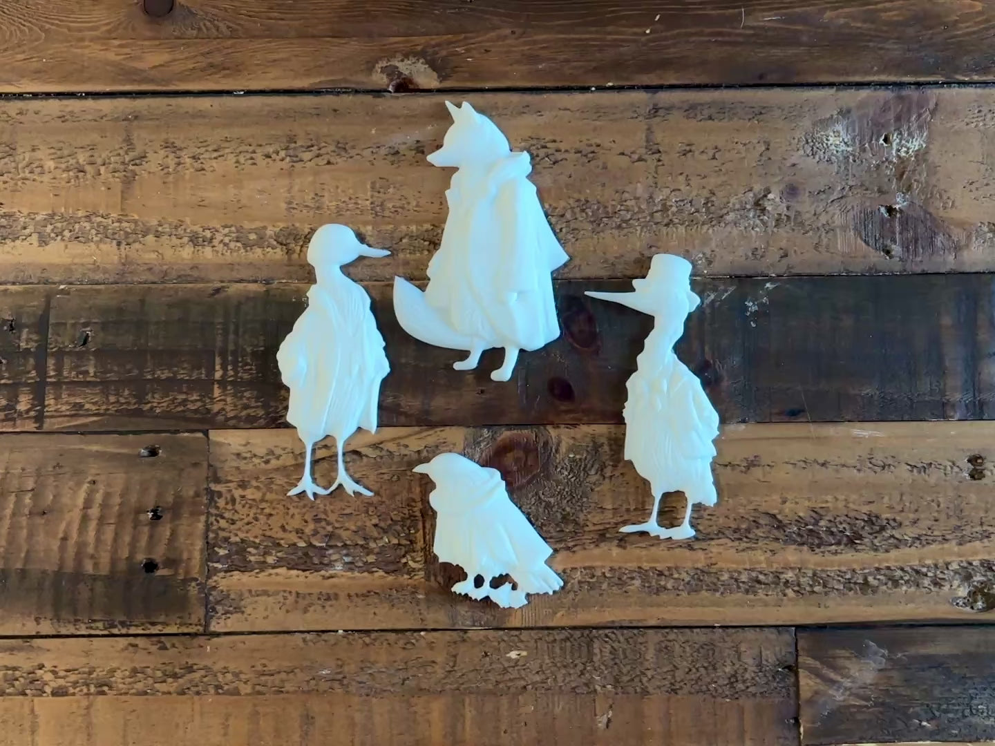 A 15 second video shows a close-up of 4 white resin castings of a fox, bird, stork, and goose all in vintage overcoats against a wood background. A hand is shown picking up the fox casting.