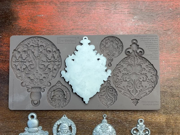 A 14 second video shows a close-up of a brown silicone mold and silver colored castings of 6 varying size ornate ornaments against a dark wood background. A hand is shown picking up and holding a middle size casting.