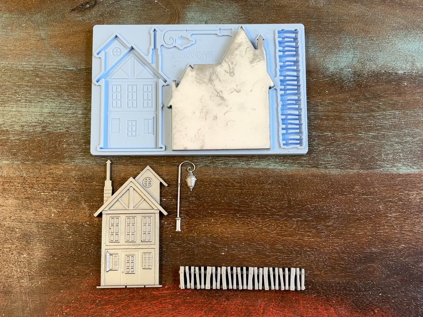 A 14 second video of LaBlanche's Houses 2 silicone mold and silver colored castings are against a wood background. A hand is shown holding one of the house castings.