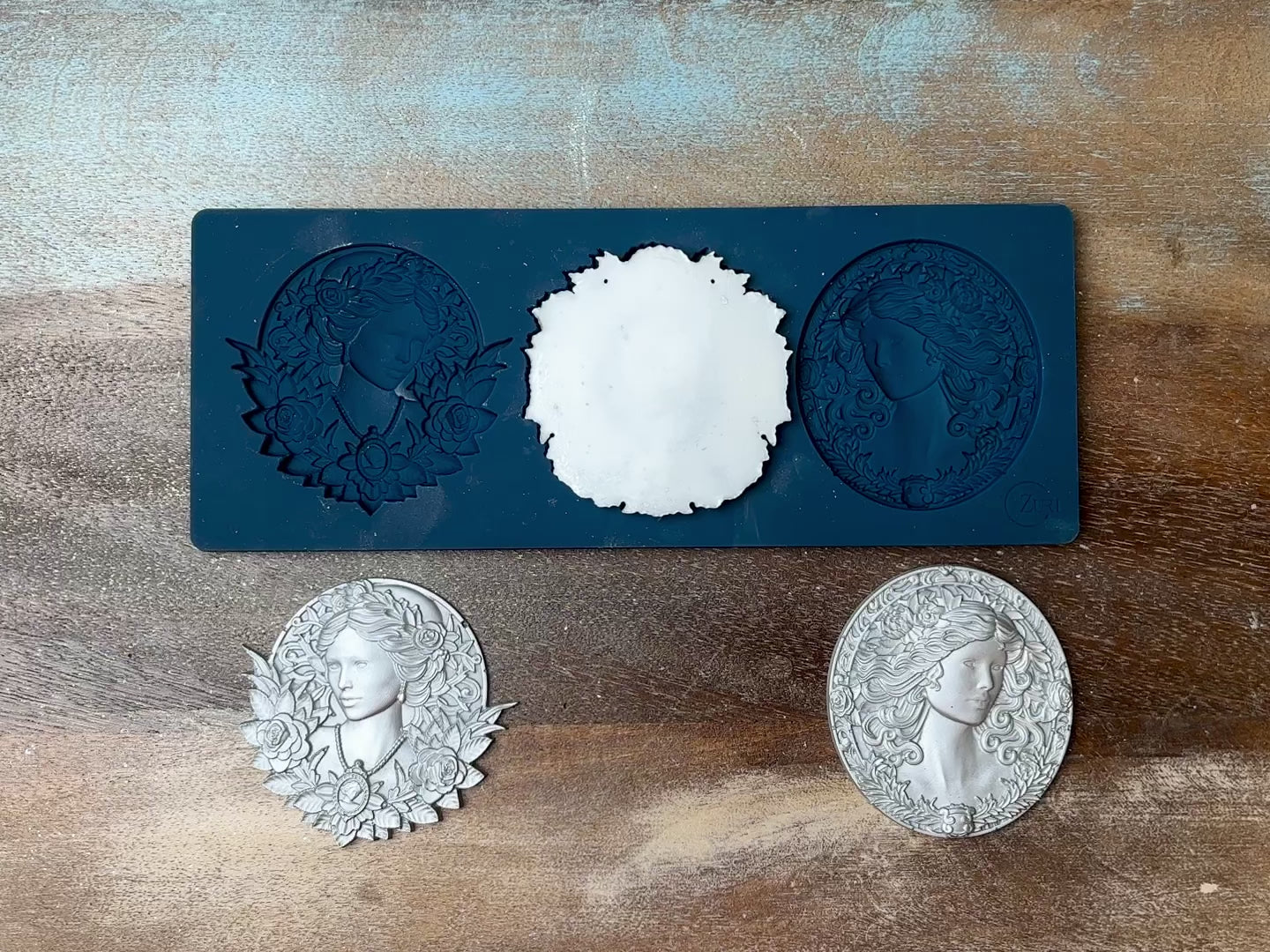 A 13 second video shows a close-up of Zuri Design's Cameo Charm silicone mold and silver colored castings against a dark wood background. A hand is shown picking up and holding one of the castings.