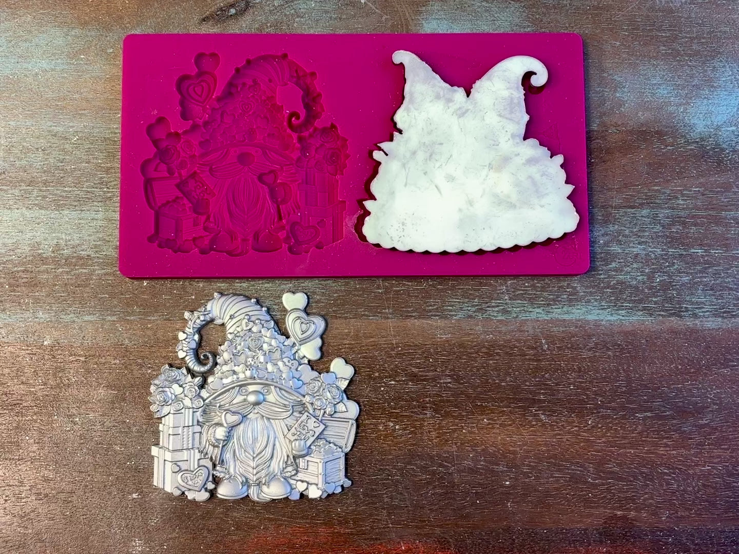 A 13 second video of Zuri Design's Valentine's Gnomes Set 1 silicone mold and silver colored castings are against a wood background. A hand is shown holding one of the castings.