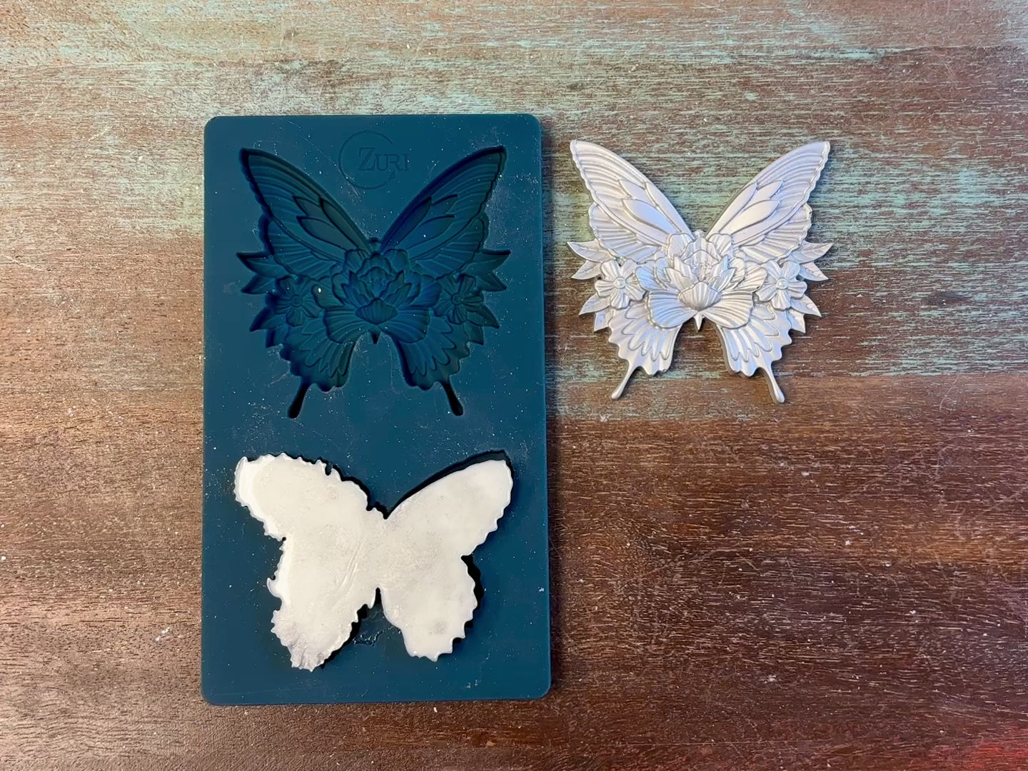 A 13 second video of Zuri Design's Butterfly Dance silicone mold and silver colored castings are against a wood background. A hand is shown holding one of the butterfly castings.