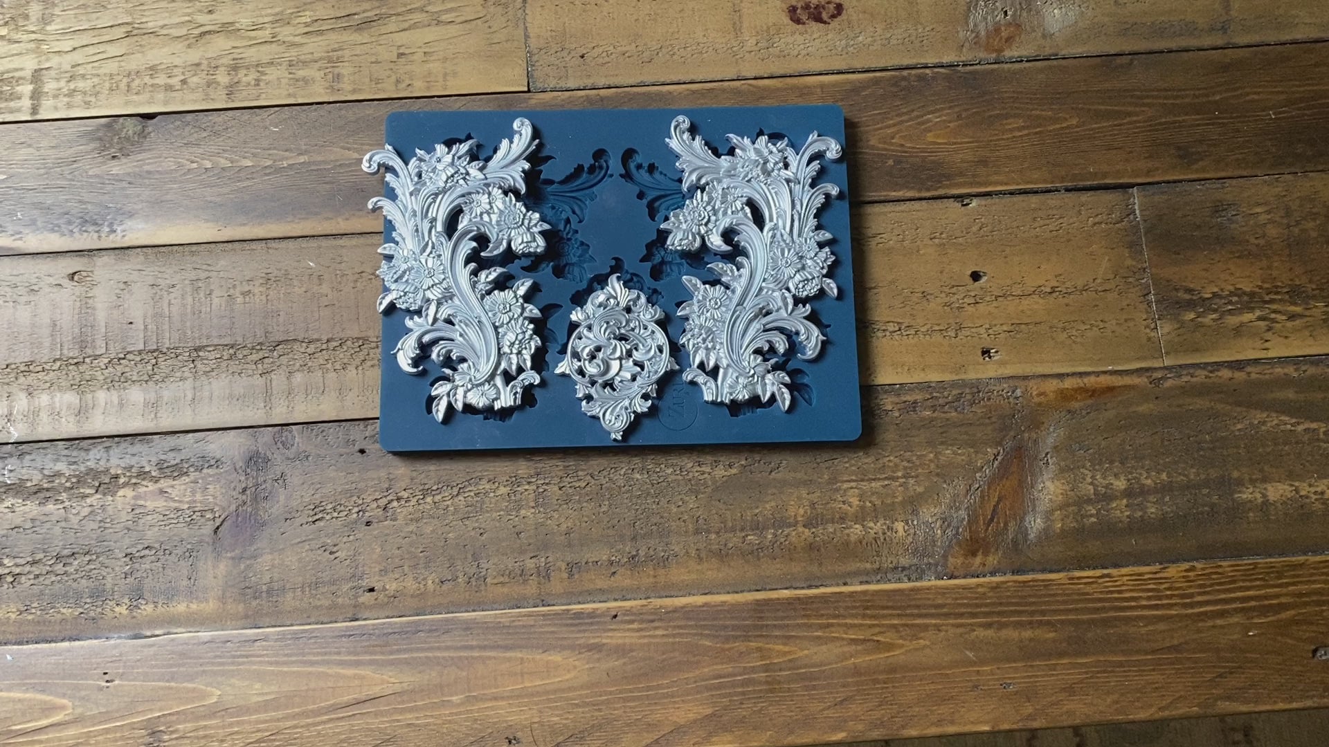 A 15 second video of Zuri Design's Flourishes Set 1 Large is against a wood background. A hand is shown holding one of the castings.