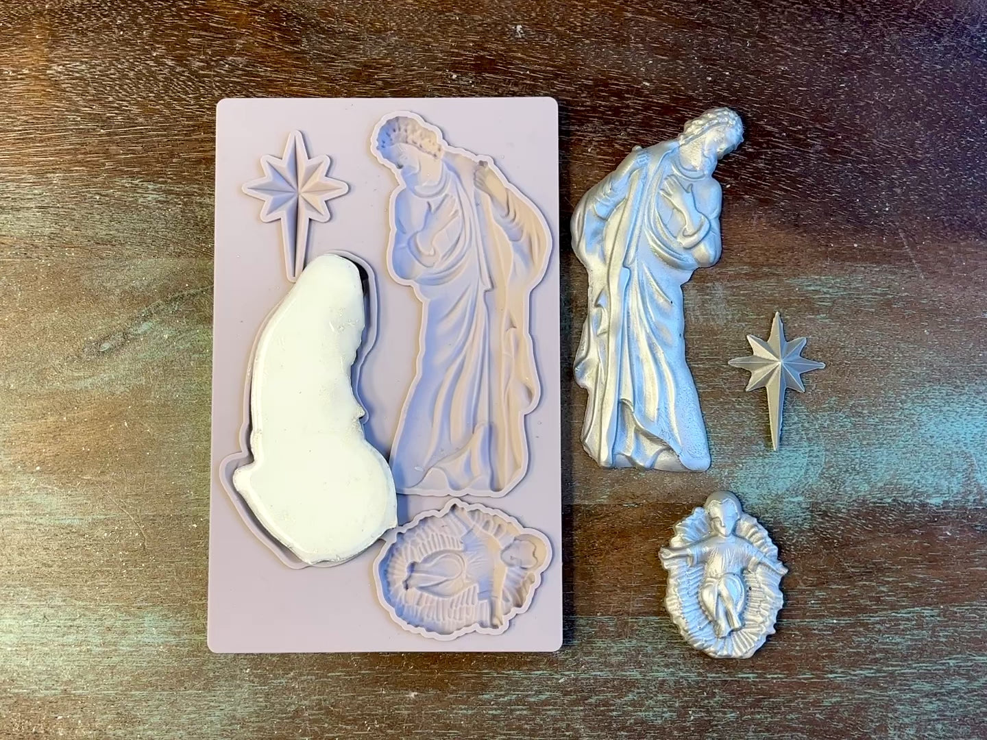 A 14 second video shows a close-up of Decoupage Queen's Holy Family silicone mold and silver colored castings against a wood background. A hand is shown holding the Mary casting.