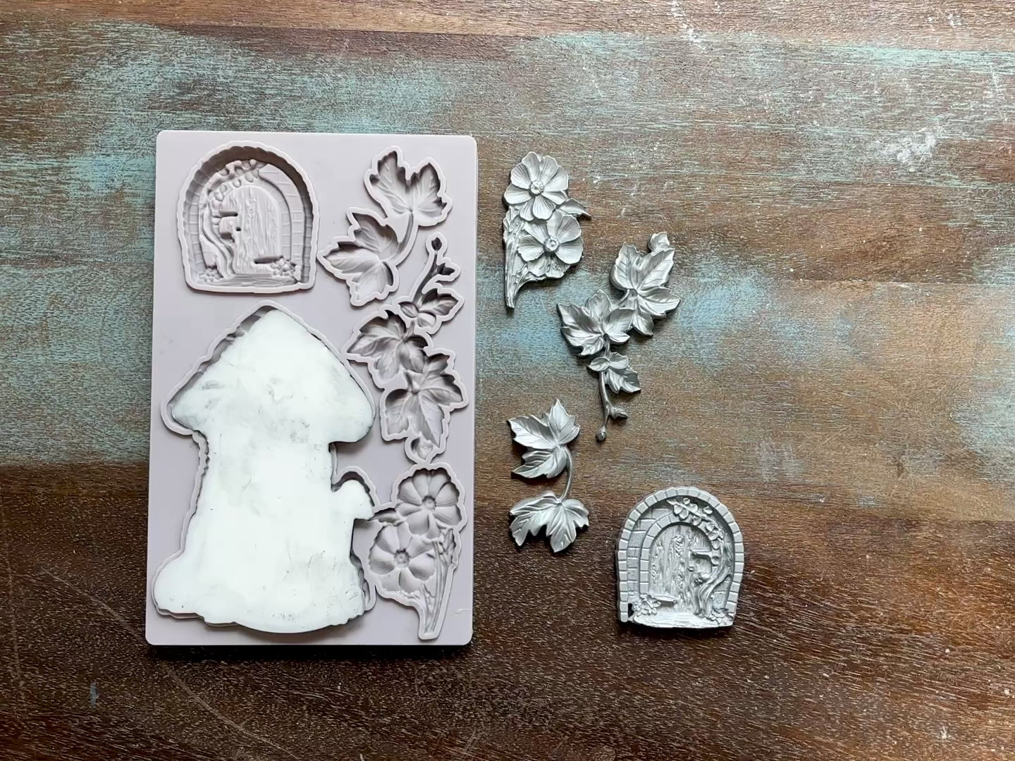 A 14 second video shows a close-up of Decoupage Queen's Toadstool House silicone mold and silver colored castings against a wood background. A hand is shown picking up the casting of the mushroom house from the mold.