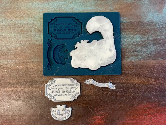 A 13 second video of Zuri Design's Cheshire Cat silicone mold and silver colored castings are against a wood background. A hand is shown holding the cat casting.
