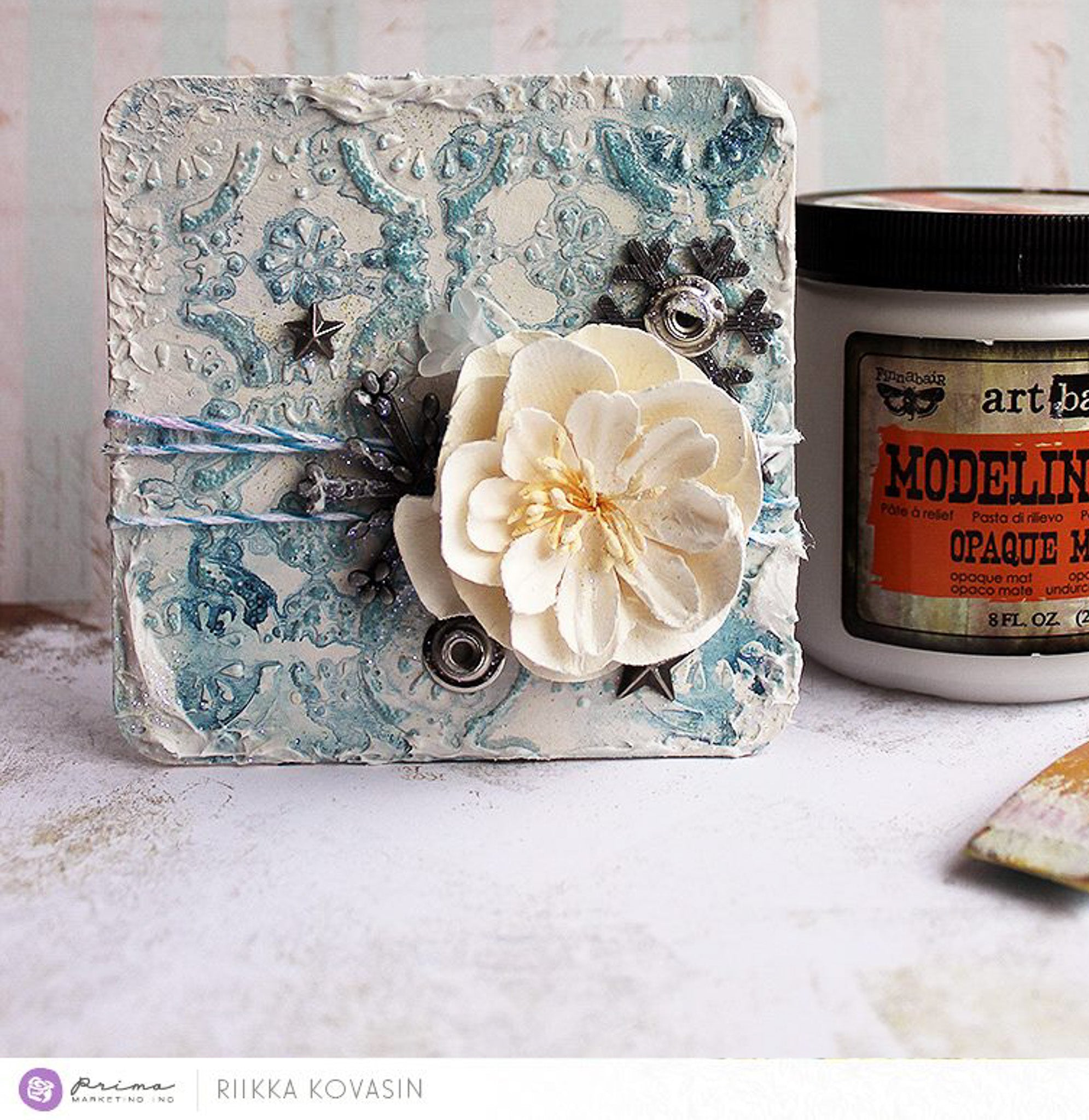 A square coaster size decor features Finnabair's Modeling Paste stenciled on it with blue watercolor and a white flower. A container of the modeling paste is to the right of the decor.