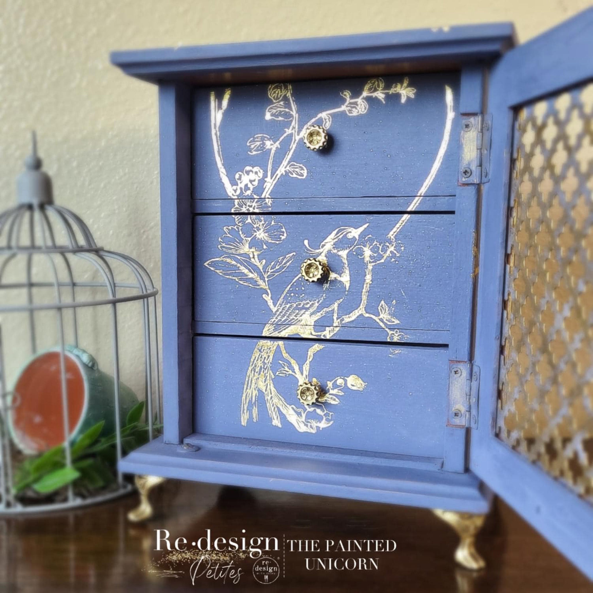 A small table-top jewelry box refurbished by The Painted Unicorn is painted a soft blue and features ReDesign with Prima's Kacha's A Bird Song Gold Foil transfer on it.