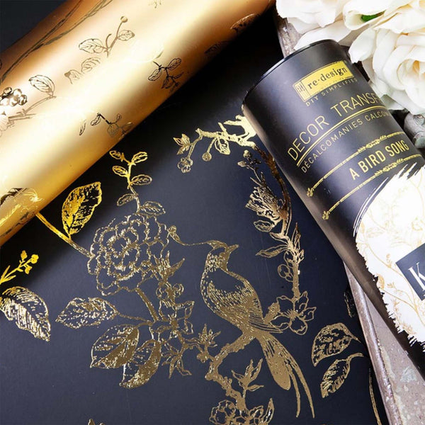 An example of Kacha A Bird Song Gold Foil is on a black surface. To the top of the example is the gold roll of the transfer. To the right is the tube of the transfer.