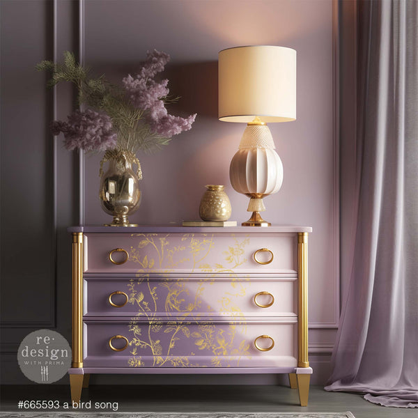 A 3-drawer dresser refurbished by ReDesign with Prima is painted light purple and cream with gold accents and features A Bird Song on its drawers.