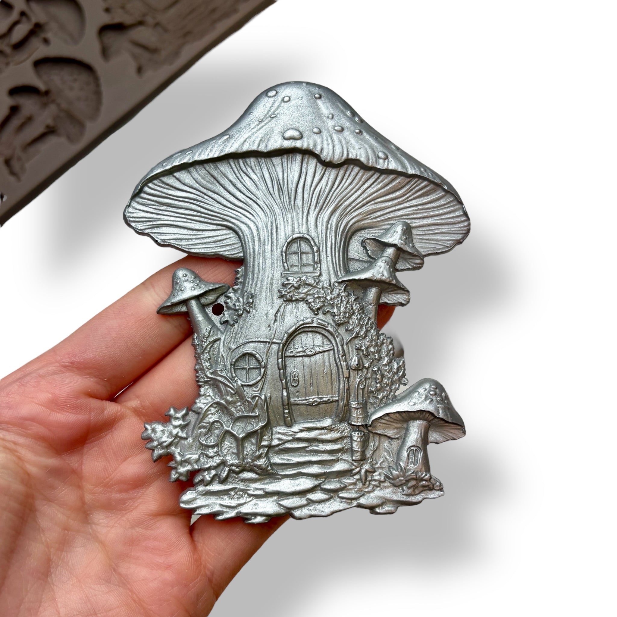 A hand is shown holding a silver colored silicone mold casting of a fairy mushroom house against a white background. A part of a brown silicone mold is visible in the background.