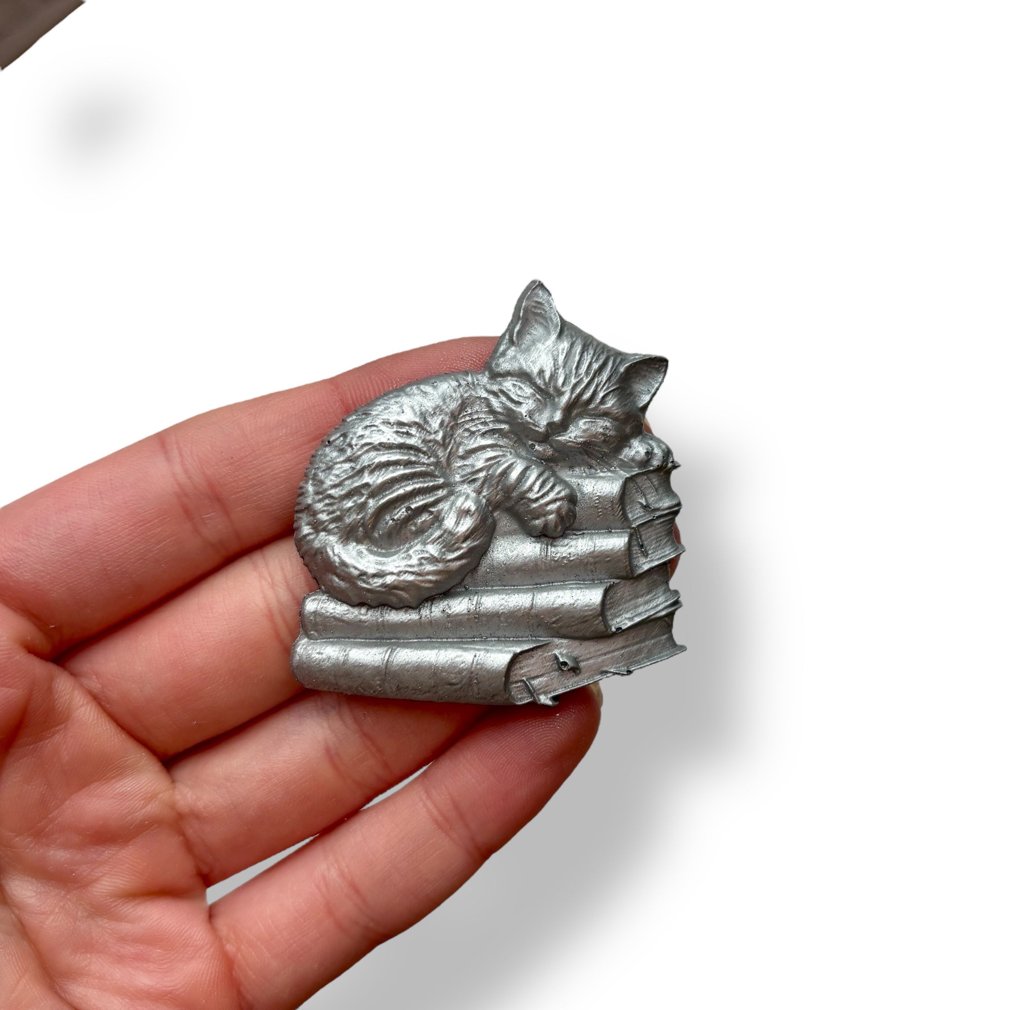 A hand is shown holding a silver colored silicone mold casting of a kitten sleeping on a stack of books against a white background.