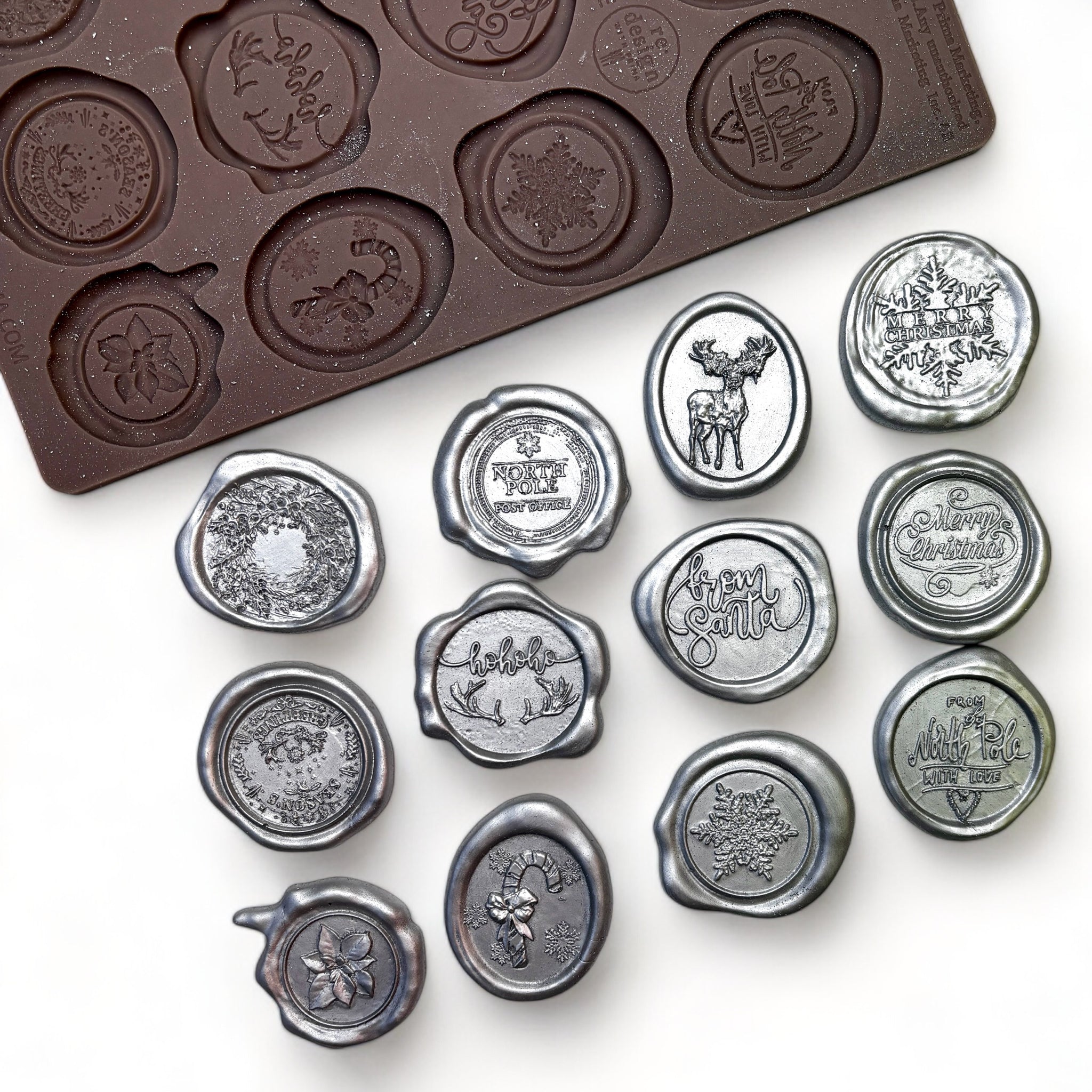 A brown silicone mold and silver colored castings of 12 wax seals that feature holiday sentiments are against a white background.