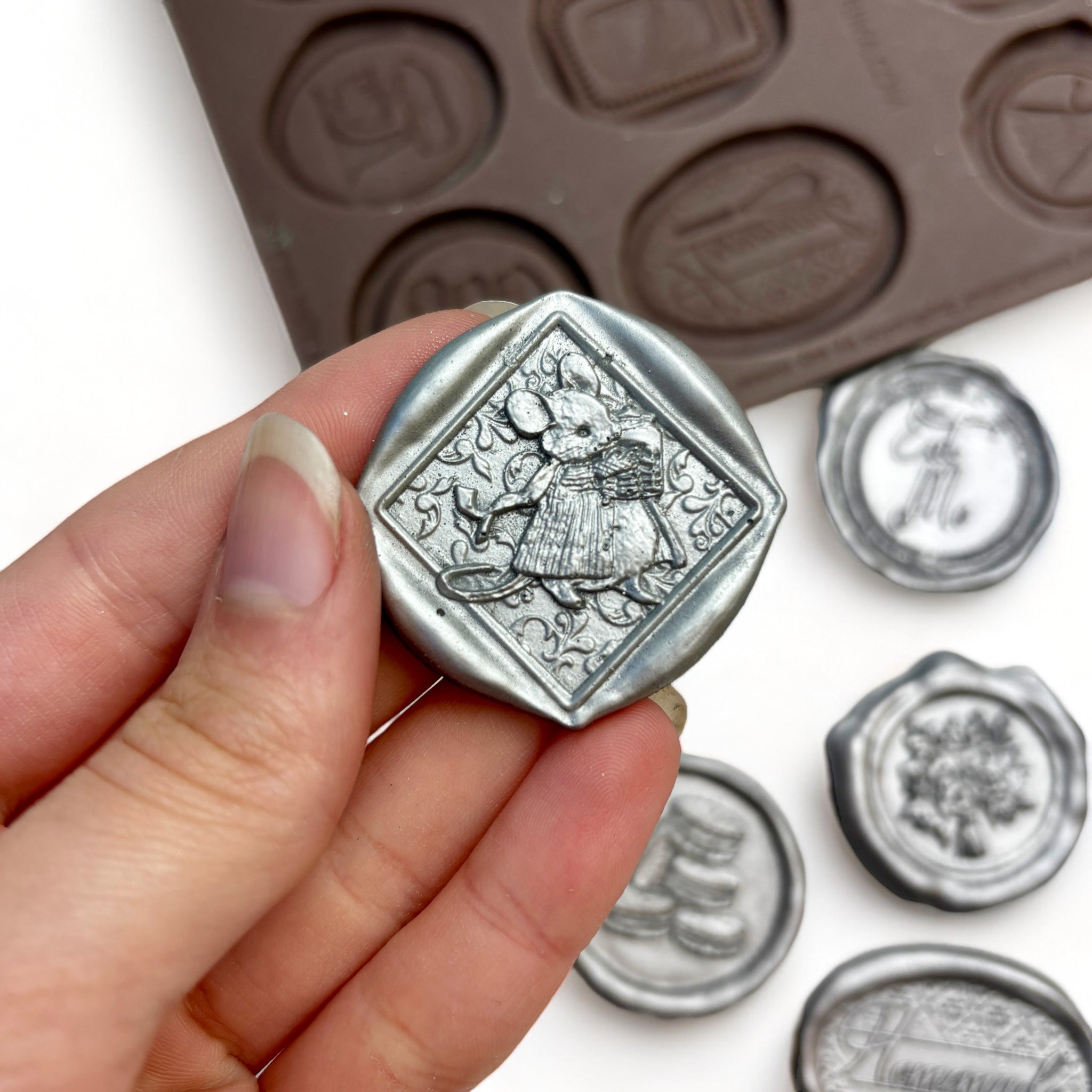 A brown silicone mold and silver colored castings of 12 baking themed wax seals are against a white background. A hand is shown holding one of the castings.