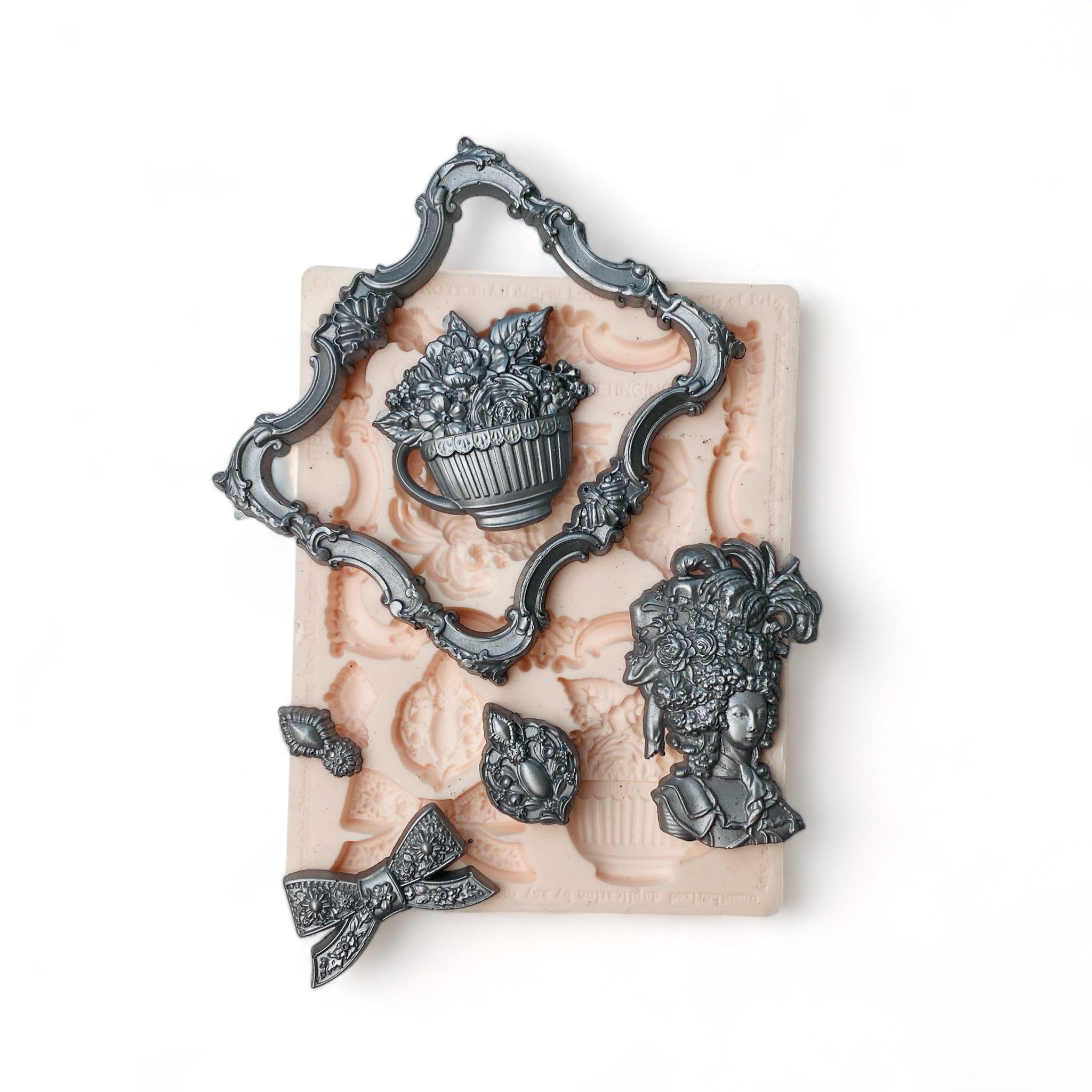 A beige silicone mold and silver colored castings of an ornate frame, ribbon bow, Marie Antoinette style head bust, bouquet of flowers in a large cup, and 2 small medallions are against a white background.