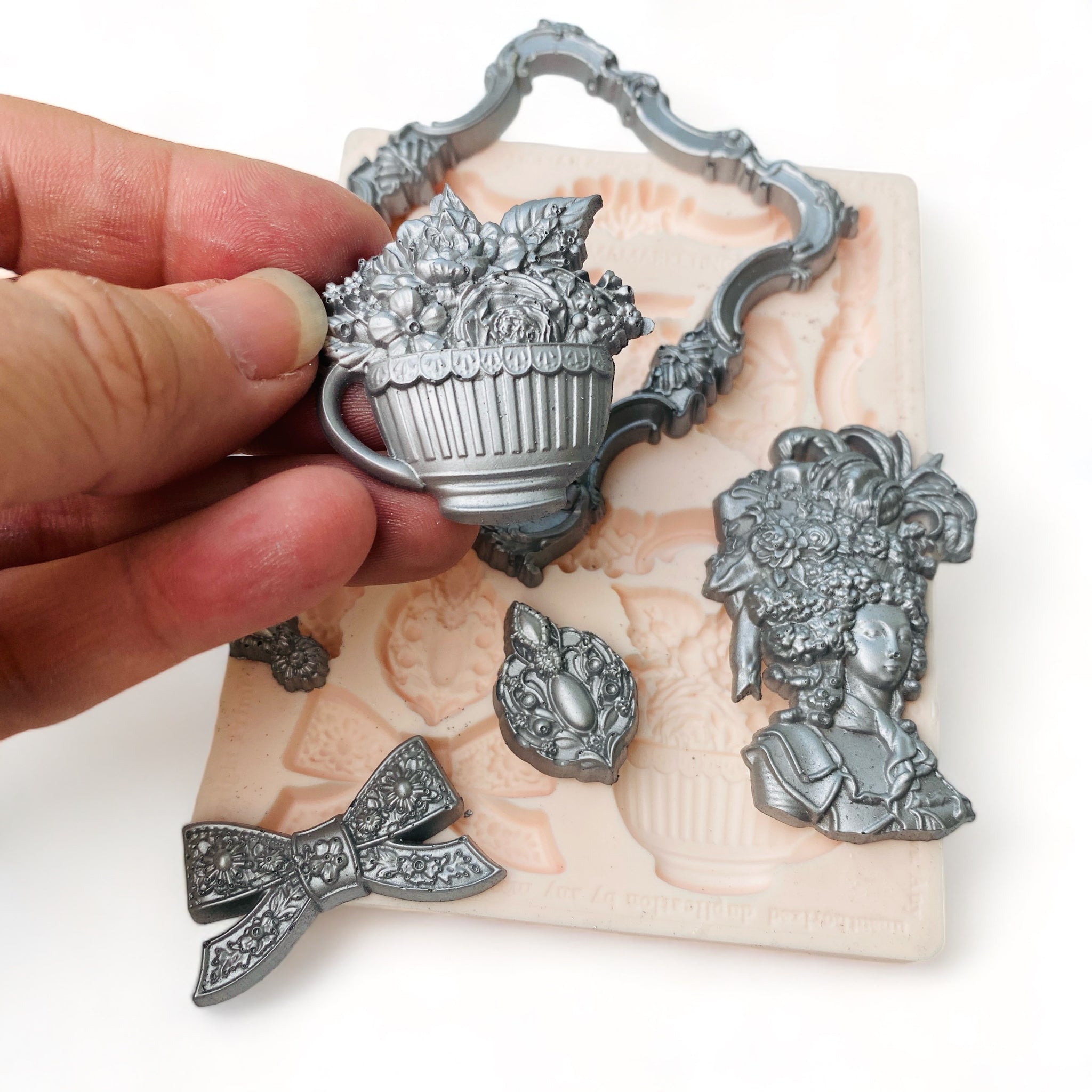 A beige silicone mold and silver colored castings of an ornate frame, ribbon bow, Marie Antoinette style head bust, bouquet of flowers in a large cup, and 2 small medallions are against a white background. A hand is shown holding the cup of flowers.