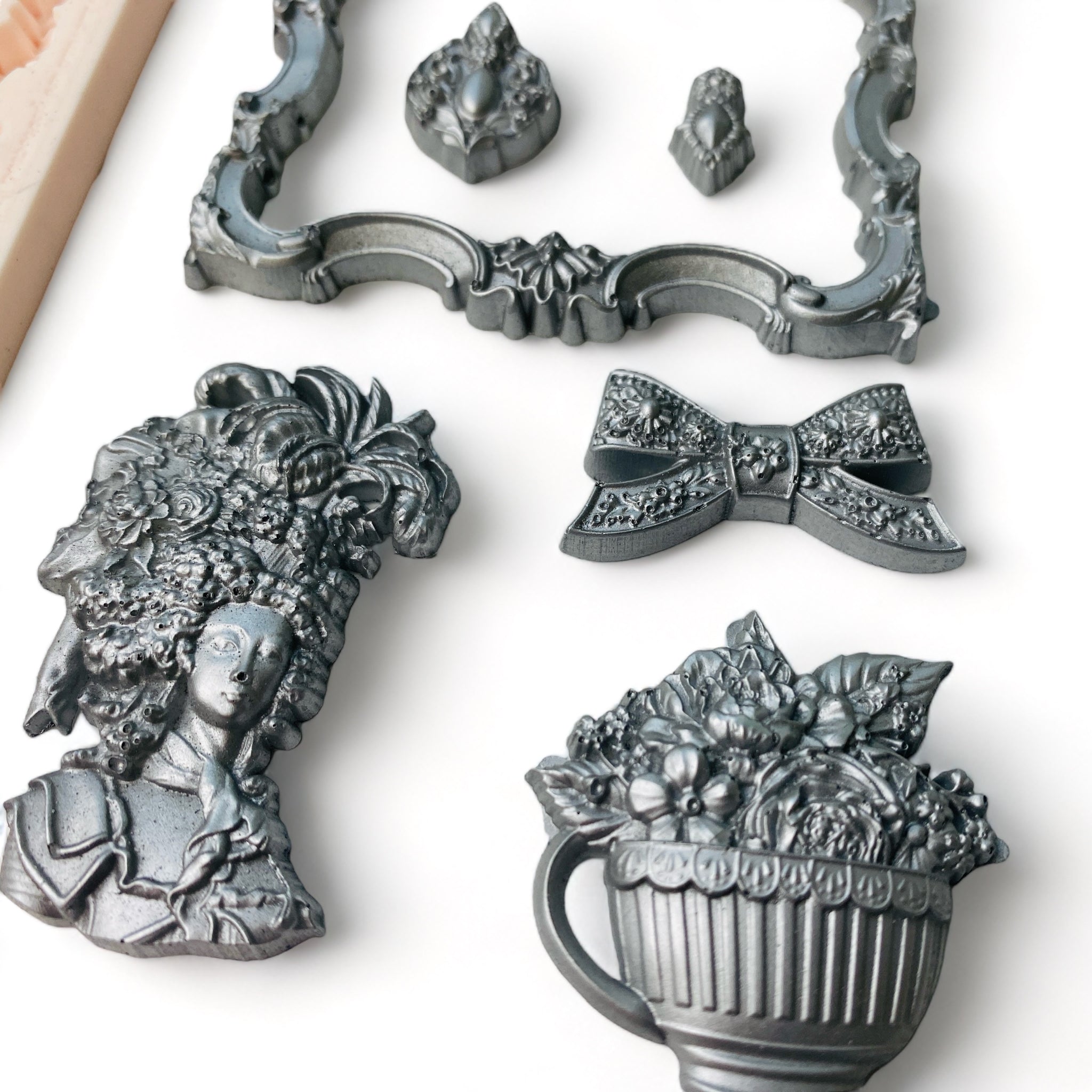 A close-up of silver colored castings of an ornate frame, ribbon bow, Marie Antoinette style head bust, bouquet of flowers in a large cup, and 2 small medallions are against a white background.