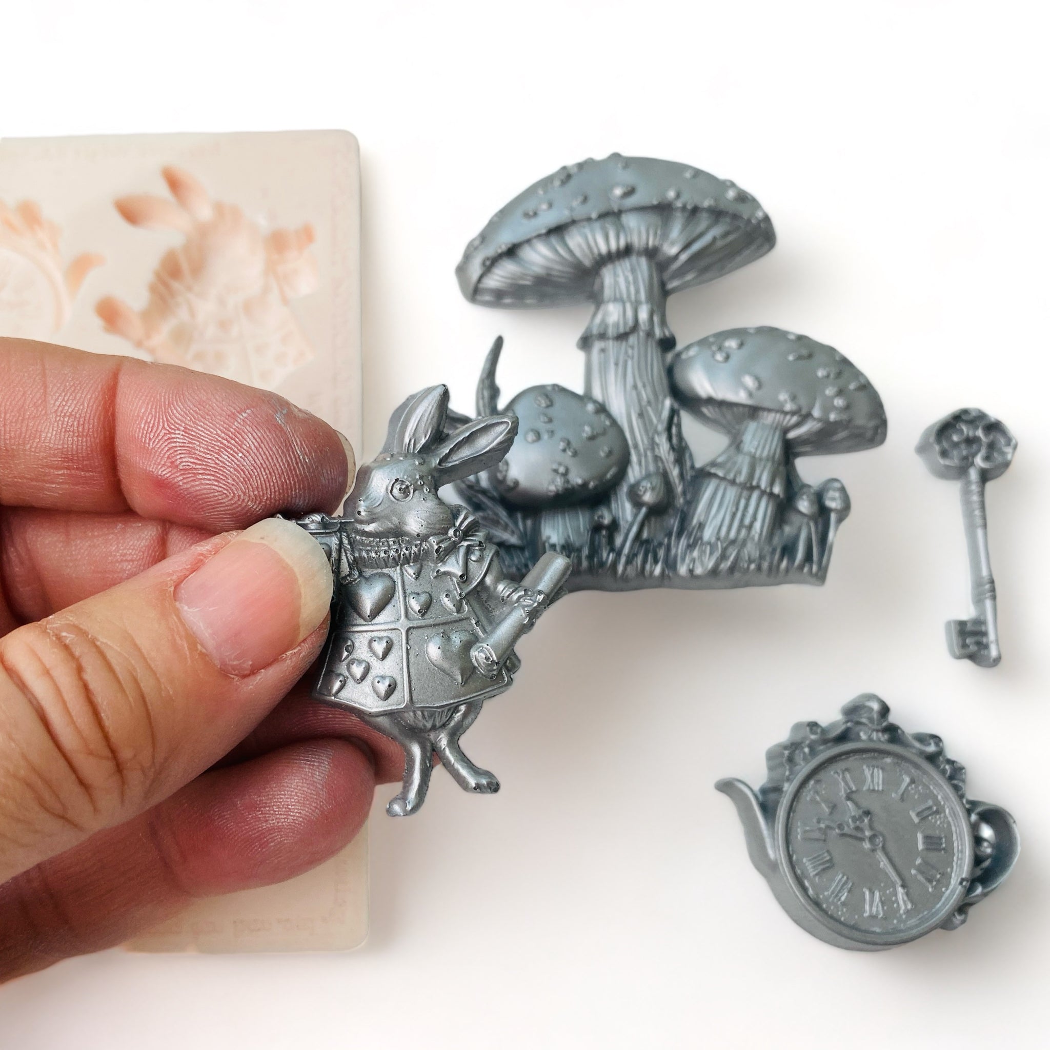 A beige silicone mold and silver colored castings of Alice in Wonderland pieces including a key, teacup clock, mushrooms, and the White Rabbit are against a white background. A hand is holding the White Rabbit casting.