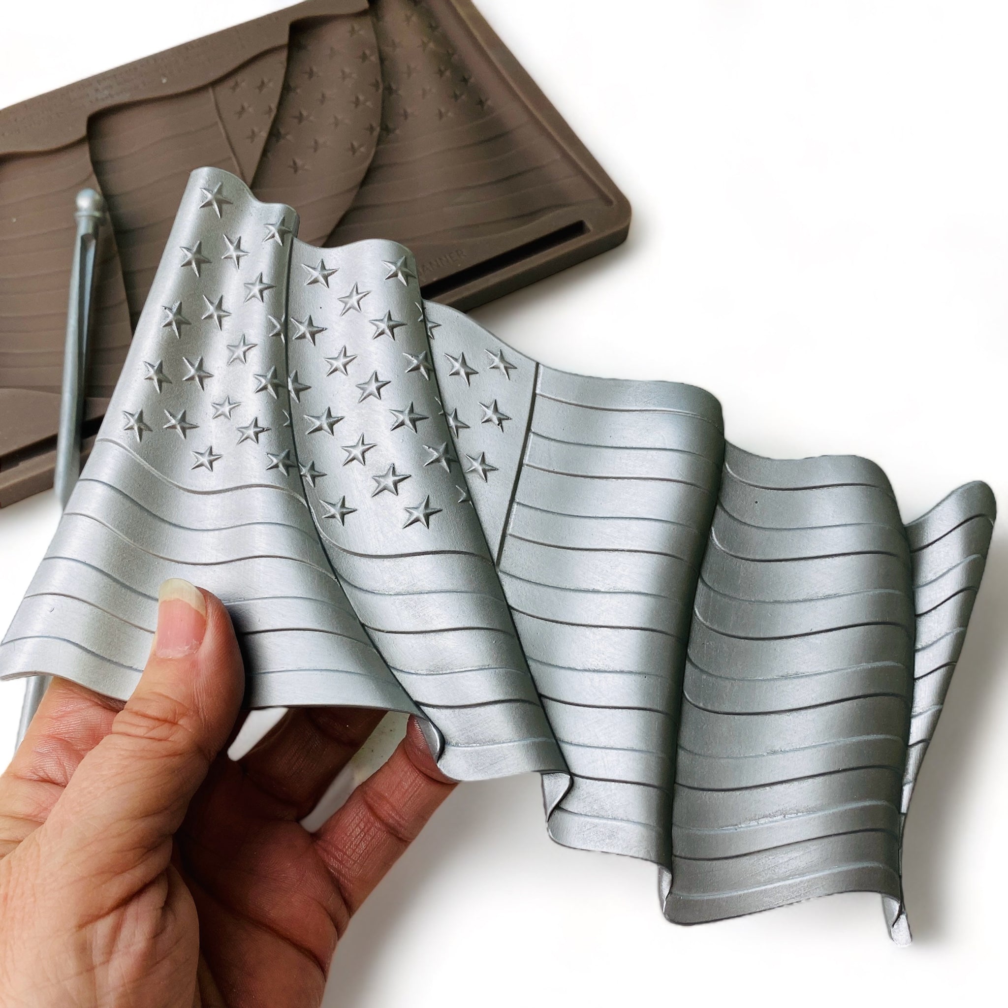 A brown silicone mold and silver colored casting of the American Flag and a flag pole are against a white background. A hand is shown holding the American flag casting.