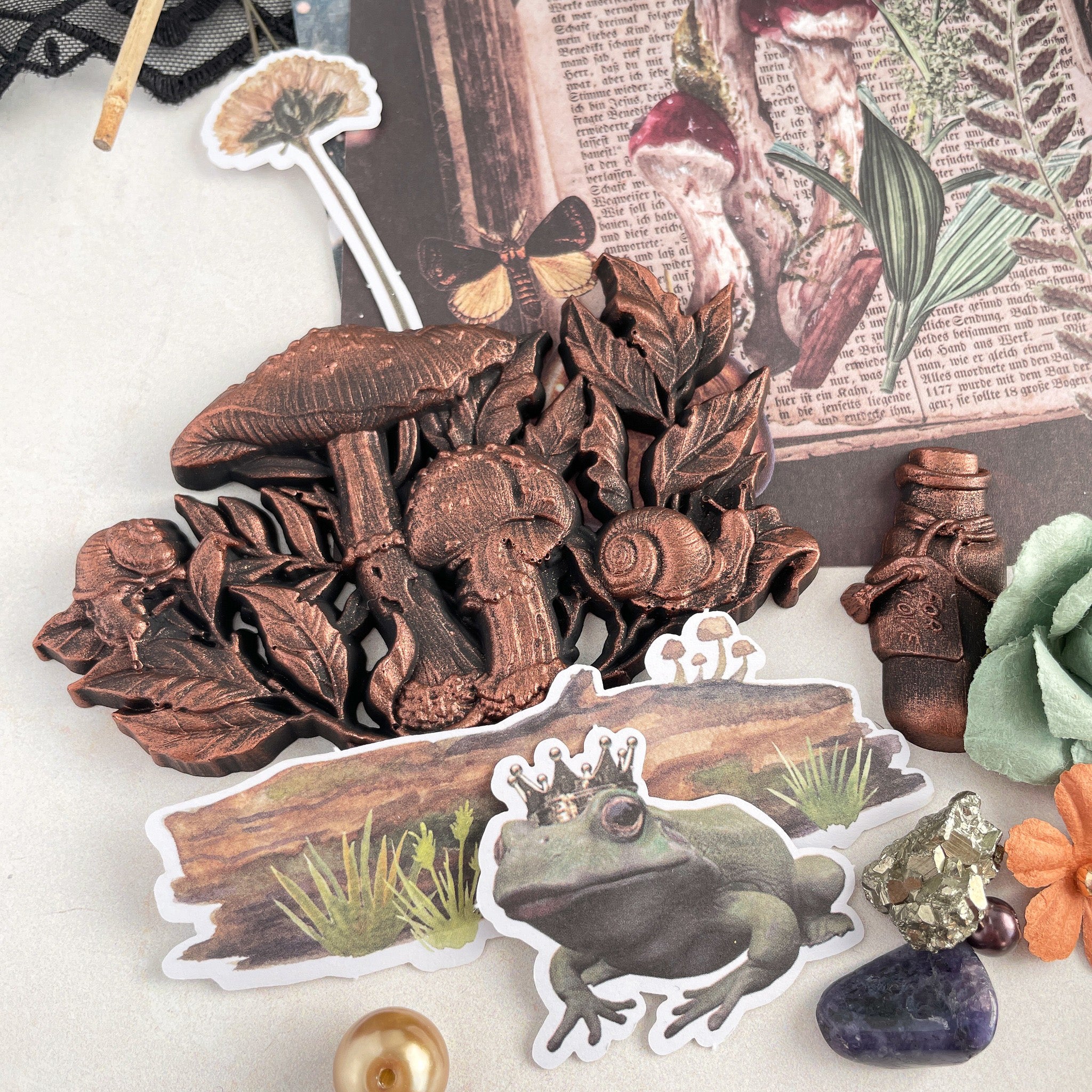 A bronze colored silicone mold casting of a cluster of mushrooms from Prima Marketing's Nature Academia silicone mold is against a white background with paper cut-outs of a frog in a crown and a log with mushrooms growing on it.