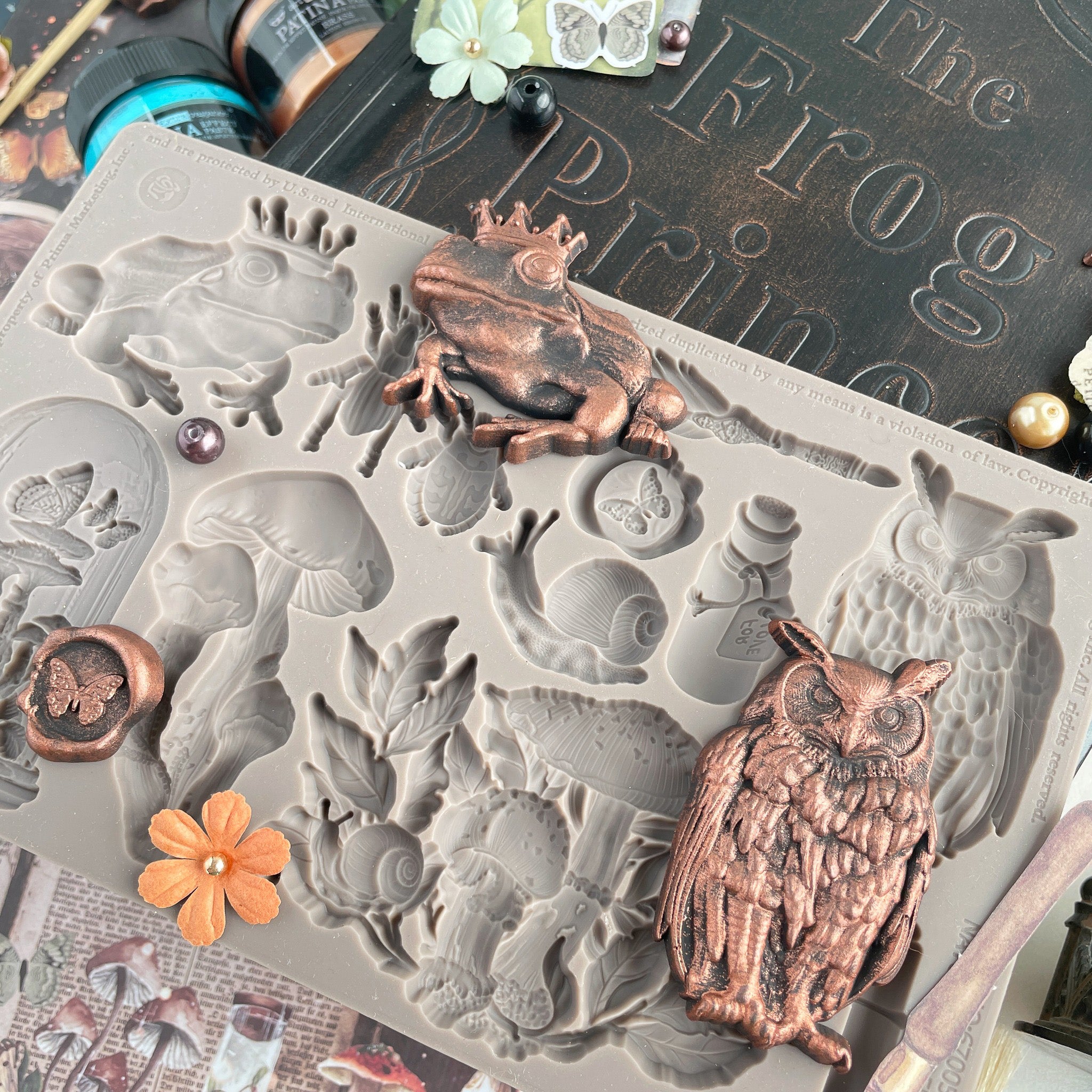 Bronze colored castings of a frog with a crown and an owl from Prima Marketing's Nature Academia silicone mold are on a project desk.