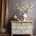 A small 4-drawer side table refurbished by Redesign with Prima is painted in a rustic white and features Rustic Patina on its drawers. A vase with flowers and some pouring pitchers sit on the table.