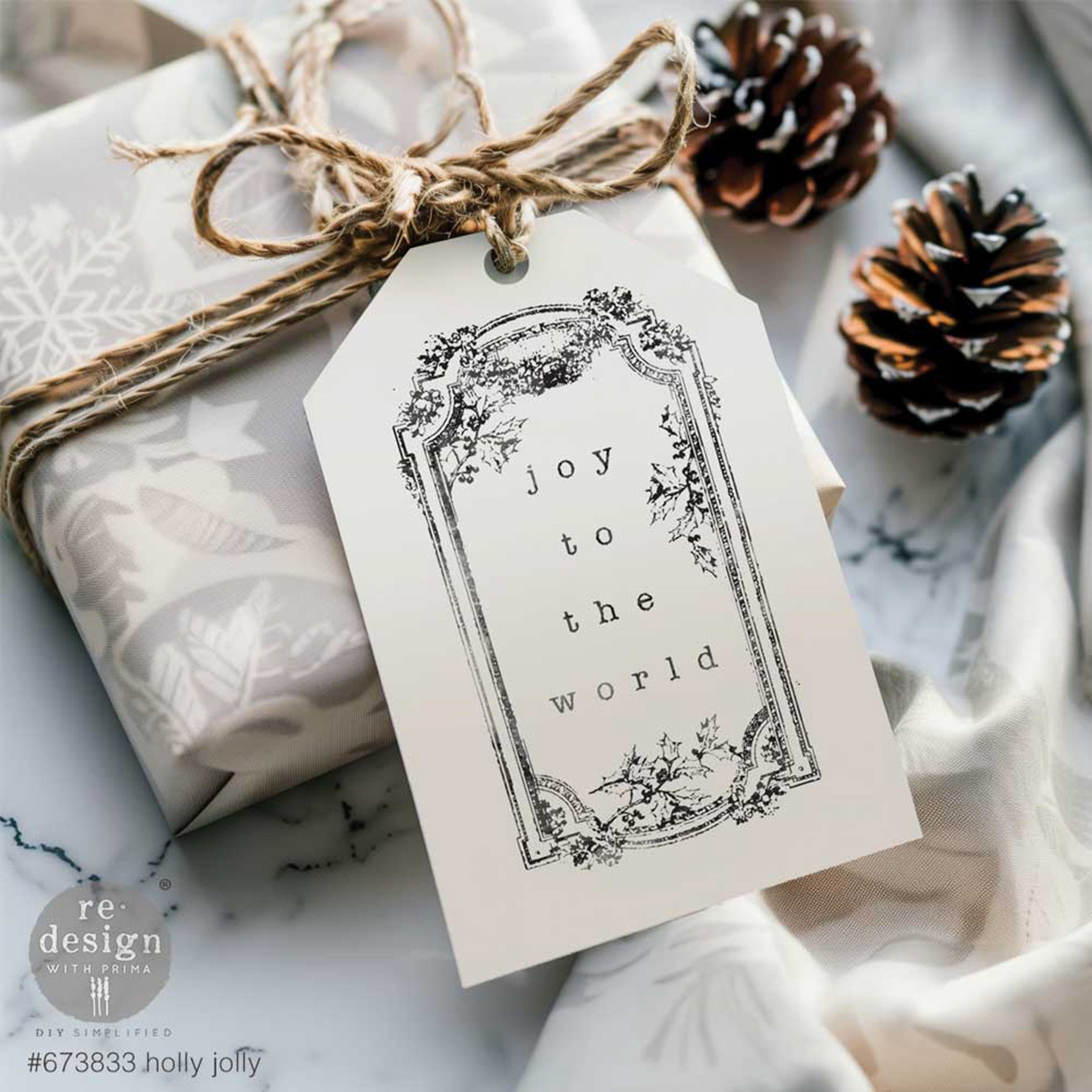 A white gift tag with a stamp that say Joy to the World created by using ReDesign with Prima's Holly Jolly decor clear stamp is on twine wrapped around a small gift with 2 pinecones to the right.