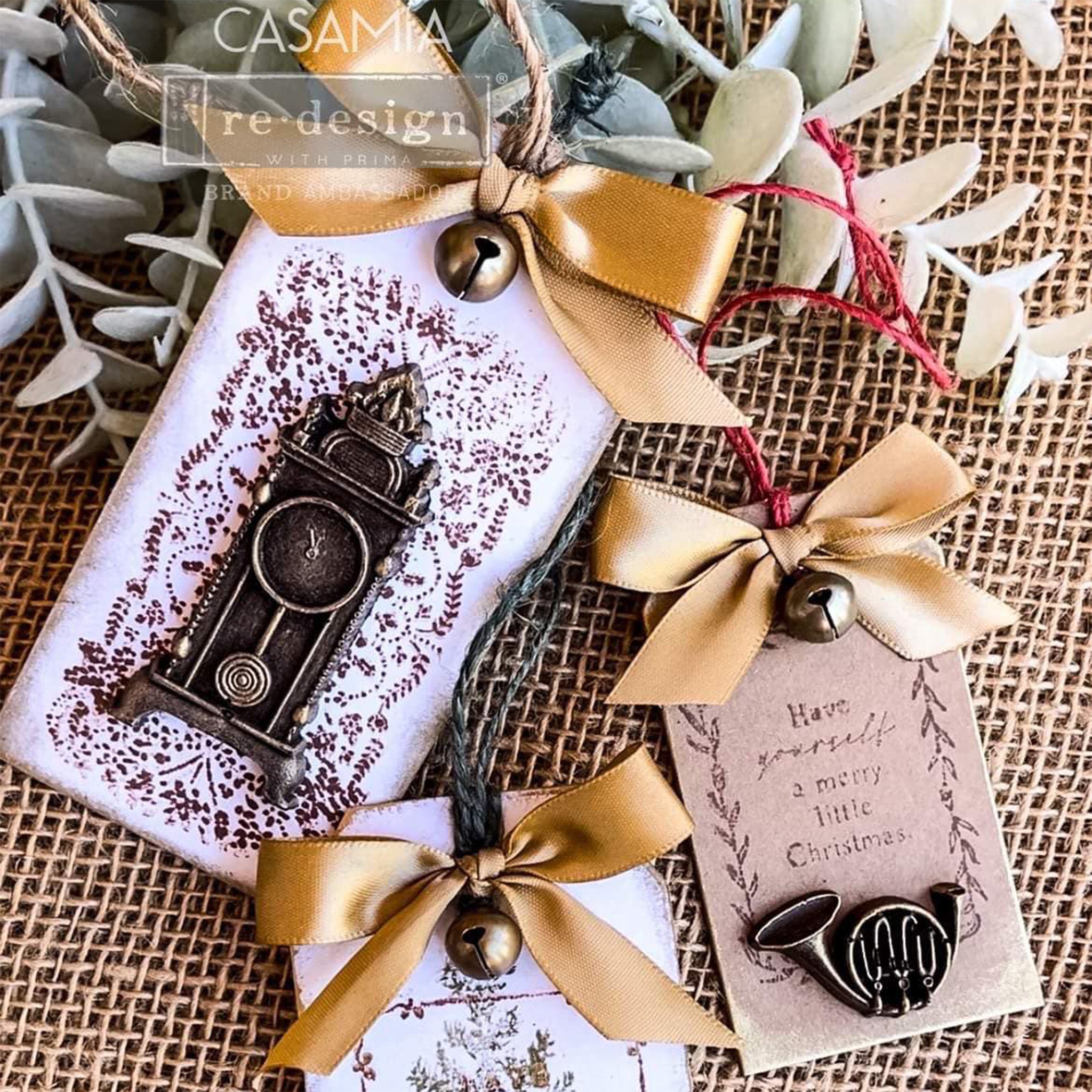 Two white and 1 brown chipboard gift tags with gold bows are against a burlap background. The brown tag features ReDesign with Prima's Merry and Bright stamp on it.