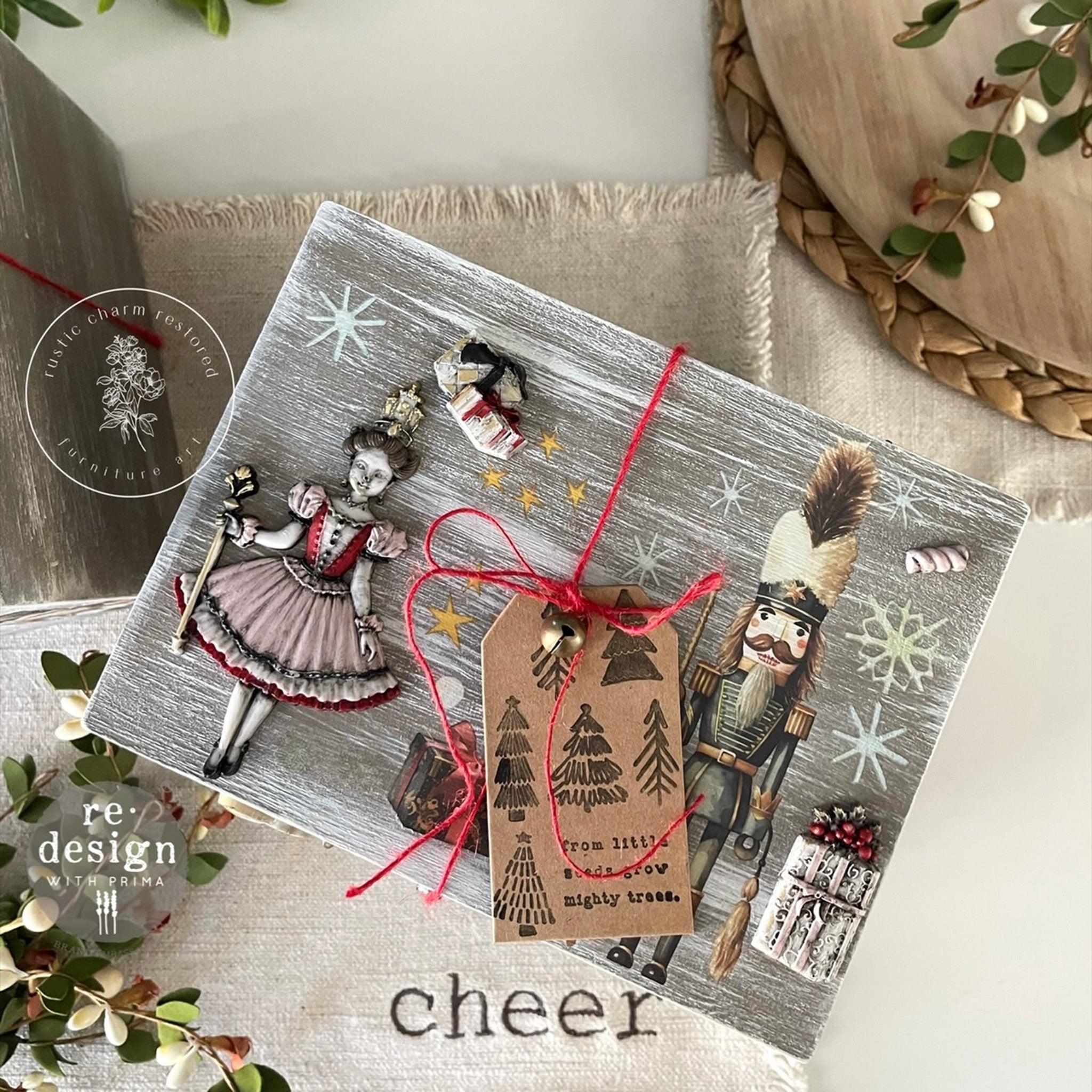 A wood craft with a nutcracker and Sugar Plum Fairy on it features a brown chipboard gift tag featuring ReDesign with Prima's Merry and Bright stamp on it.