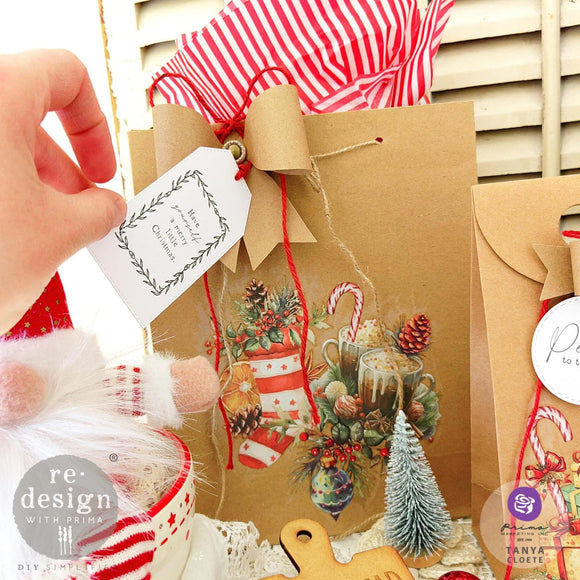 A brown paper gift bag features ReDesign with Prima's Winter Wonderland Wonders small transfers on it and has a white gift tag that features ReDesign with Prima's Merry and Bright decor stamp on it.