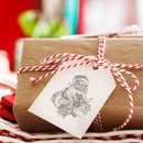 A brown paper wrapped gift has a white gift tag that features a stamp from ReDesign with Prima's Santa's Helper Decor Stamp.