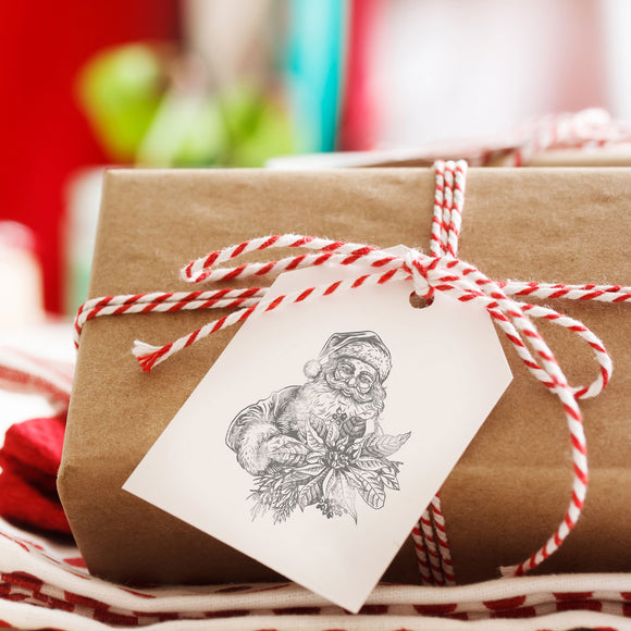 A brown paper wrapped gift has a white gift tag that features a stamp from ReDesign with Prima's Santa's Helper Decor Stamp.