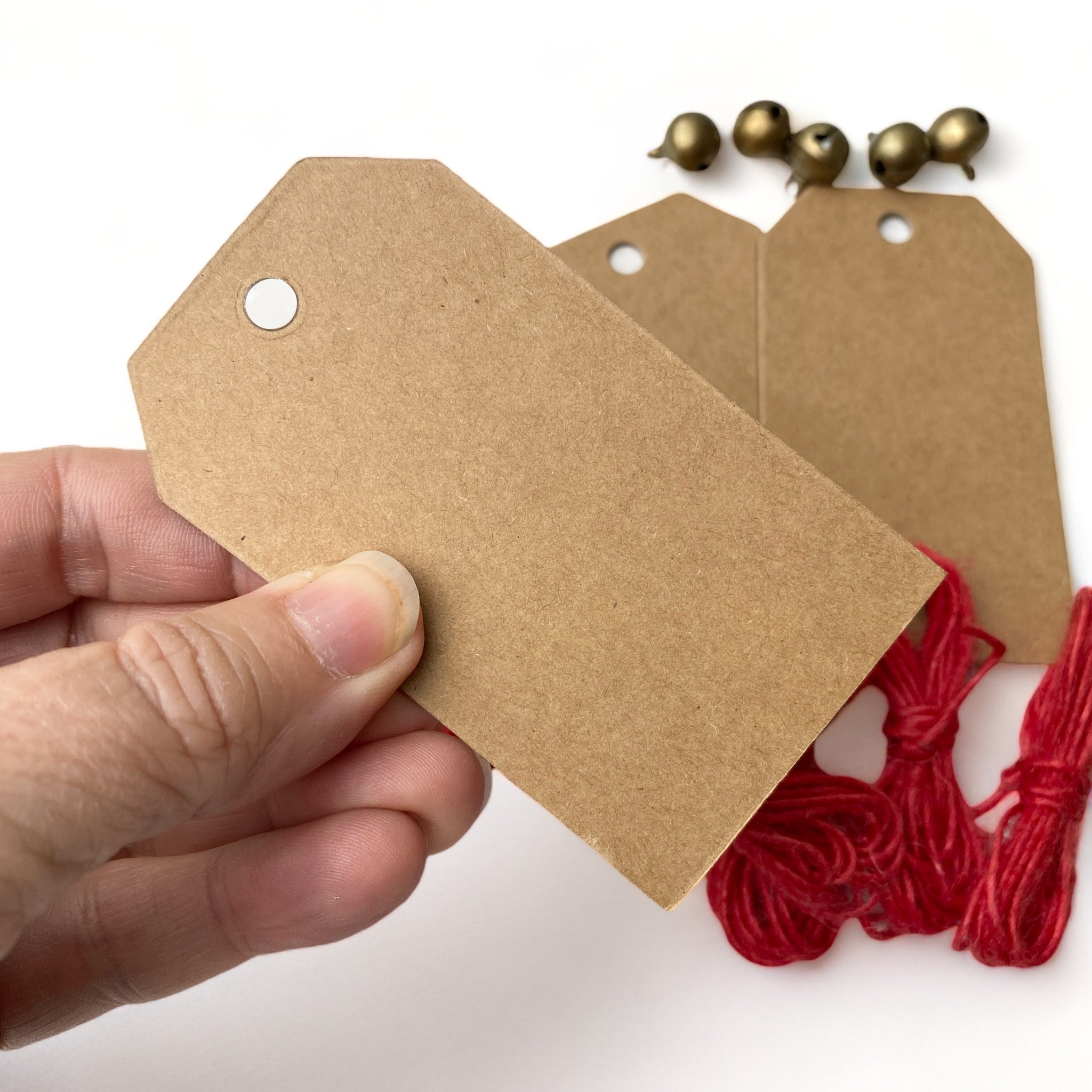 A hand is shown holding a brown chipboard Christmas gift tag. Behind it are more gift tags, small round gold bells, and red string all against a white background.