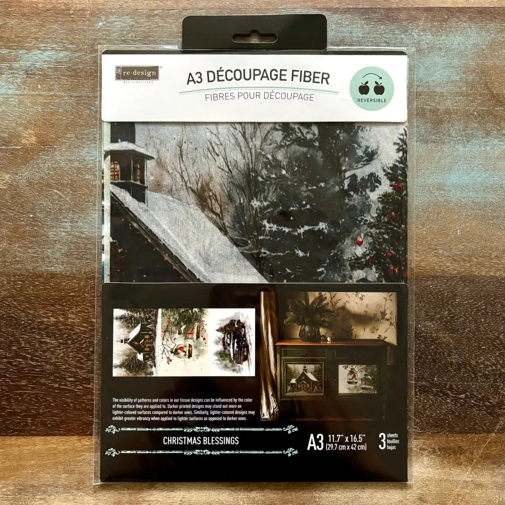 A package of ReDesign with Prima's Christmas Blessings A3 fiber paper 3 pack is against a wood background.