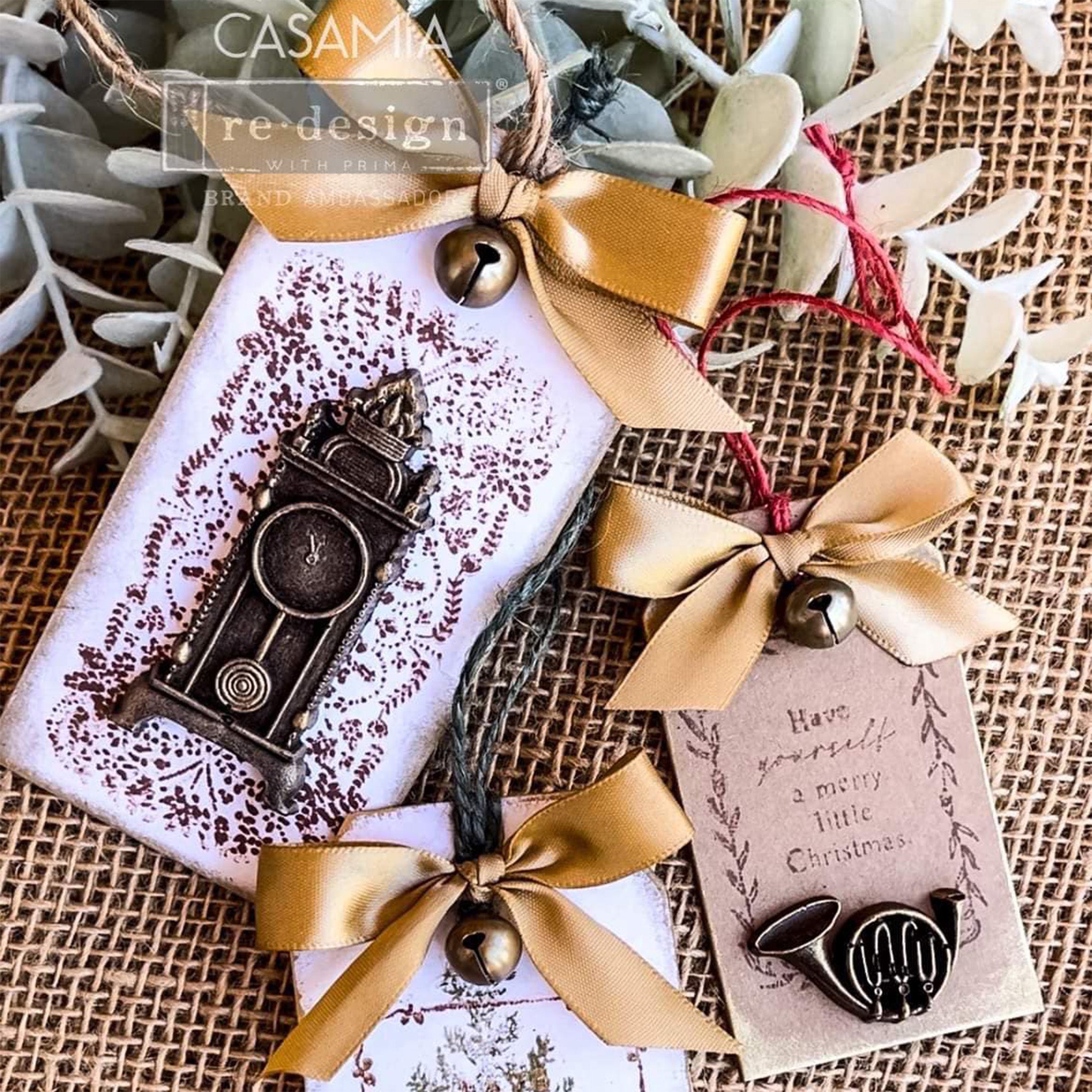 A close-up of 2 white and 1 brown tag created from ReDesign with Prima's Christmas Chipboard Tags are against a burlap background.