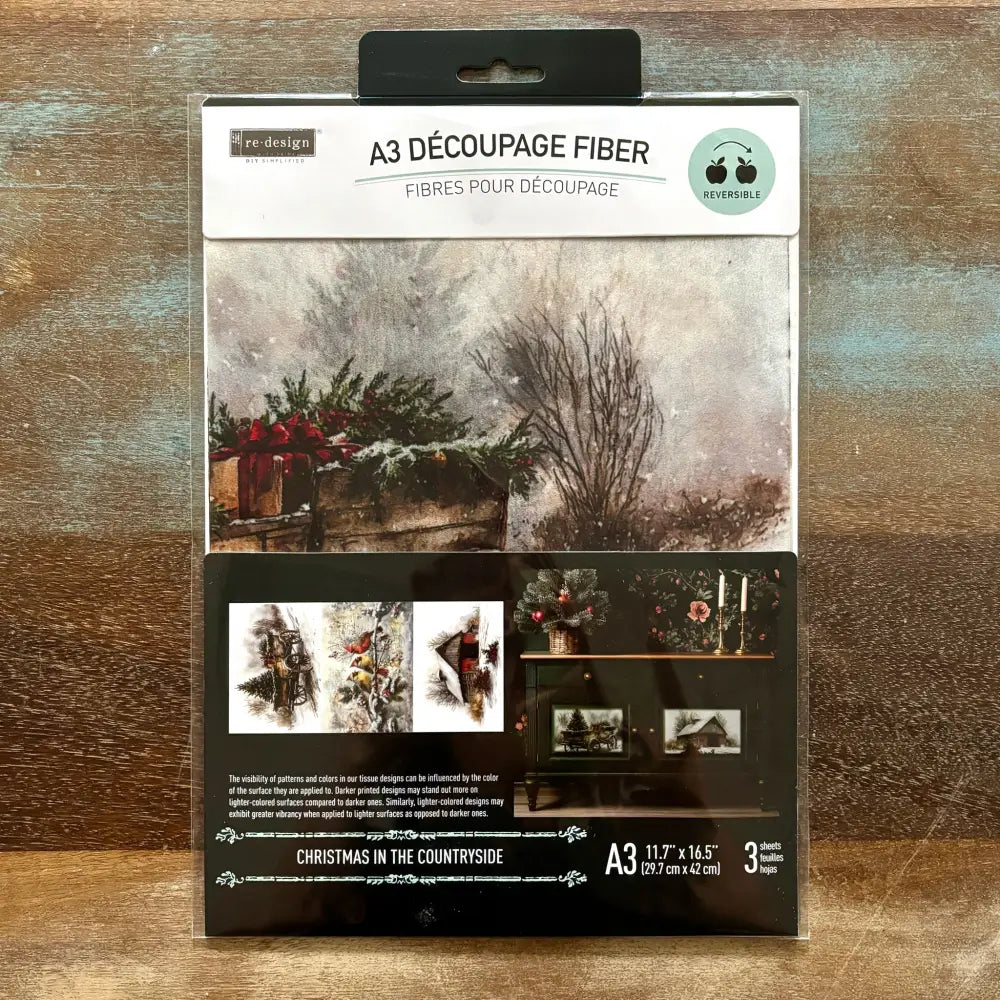 A package of ReDesign with Prima's Christmas in the Countryside A3 fiber paper 3 pack is against a wood background.