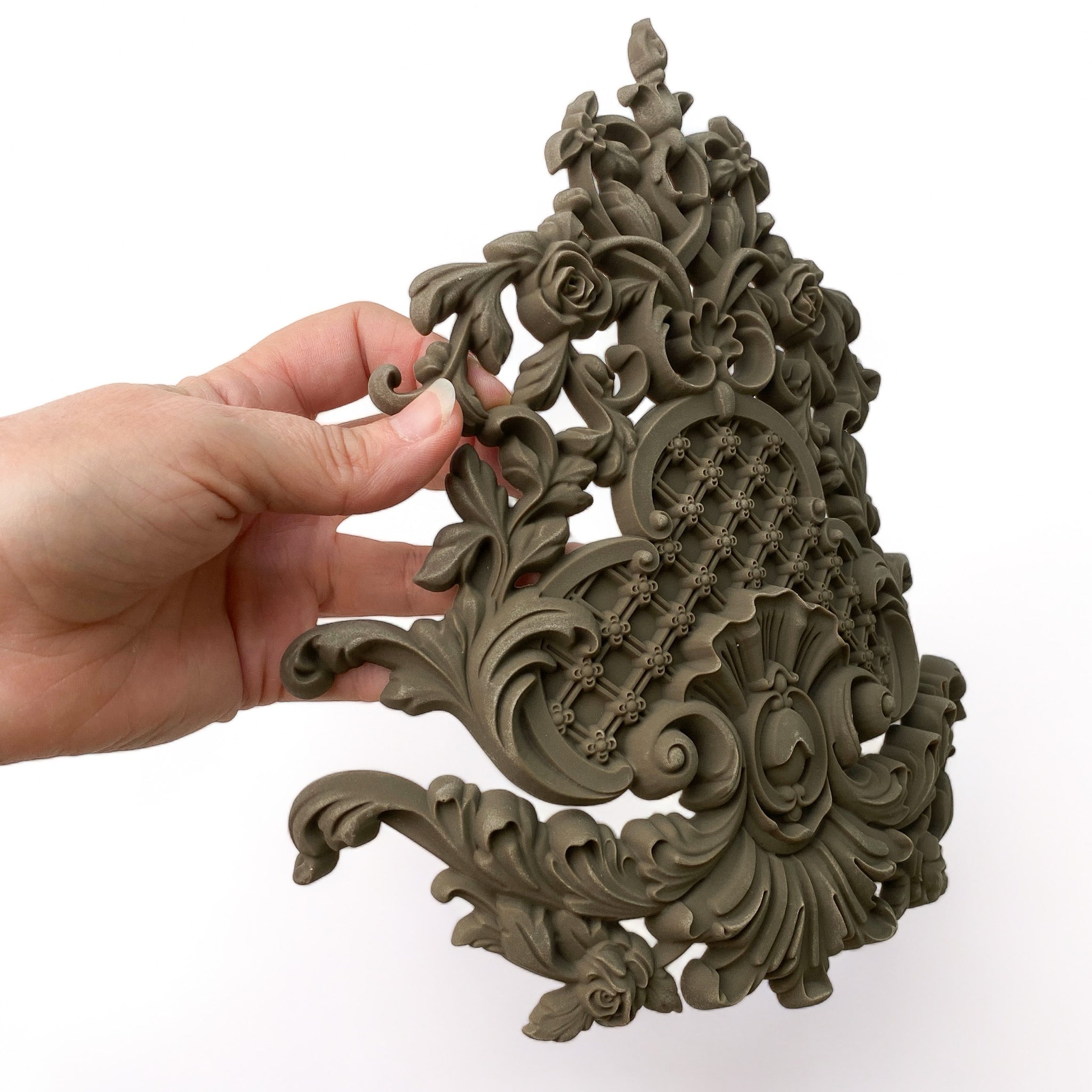 A hand is holding a curved bendable furniture applique of an intricate Baroque flourish with lattice against a white background.